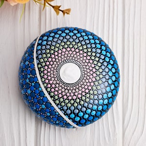 May include: A round, blue, pink, and green painted rock with a white circle in the center. The rock is decorated with a mandala pattern.