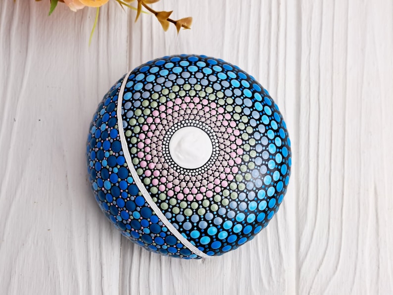 May include: A round, blue, pink, and green painted rock with a white circle in the center. The rock is decorated with a mandala pattern.