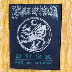 Op de afbeelding: Zwarte en witte geborduurde patch met een zilverkleurig gevleugeld wezen in een cirkel met een halve maan. De tekst "CRADLE OF FILTH" staat boven de cirkel en "DUSK and her embrace" staat onder de cirkel.