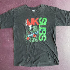May include: Black t-shirt featuring a graphic of a soldier holding a rifle, with the words "UK SUBS" in green and red. The word "warhead" is printed vertically. The shirt is made of a soft material.