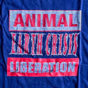 以下が含まれることがあります： 赤と白で「ANIMAL EARTH CRISIS LIBERATION」とプリントされた青いTシャツです。