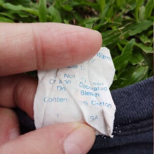 May include: A white tag with blue text that reads "Machine Wash Warm, Do Not Bleach, Do Not Iron, Decorated, 100% Cotton, Made in USA."