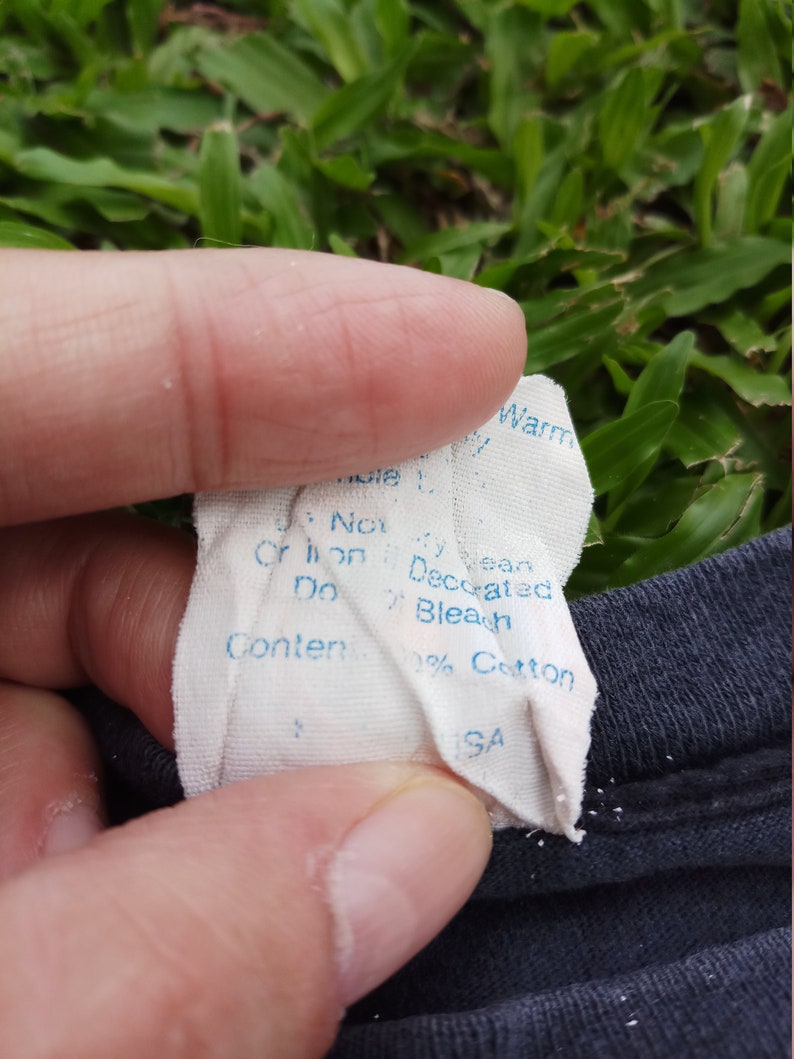 May include: A white tag with blue text that reads "Machine Wash Warm, Do Not Bleach, Do Not Iron, Decorated, 100% Cotton, Made in USA."