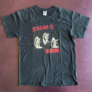 May include: Black t-shirt with the text "STALAG 13" in red above a graphic of three figures. Below the figures, the text "IN CONTROL" is also in red. The shirt is a crew neck style.