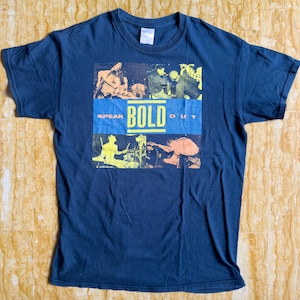 May include: A dark blue t-shirt with a graphic print of a band. The graphic features a yellow and black design with the words "Speak Bold Out".