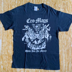 May include: Black t-shirt with a white graphic featuring the text "Cro-Mags" above a detailed illustration of a monster and figures. The text "Show You No Mercy" is below the graphic.