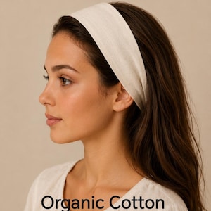 May include: A wide, off-white headband made of organic cotton fabric. The headband is worn across the forehead and holds back long brown hair. The text on the image reads Organic Cotton 35+ colours, indicating the available variety for this accessory.