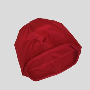 May include: A solid red beanie hat made of soft fabric. The hat is a simple, classic design, with a folded brim and a rounded crown. The hat is shown against a light gray background.