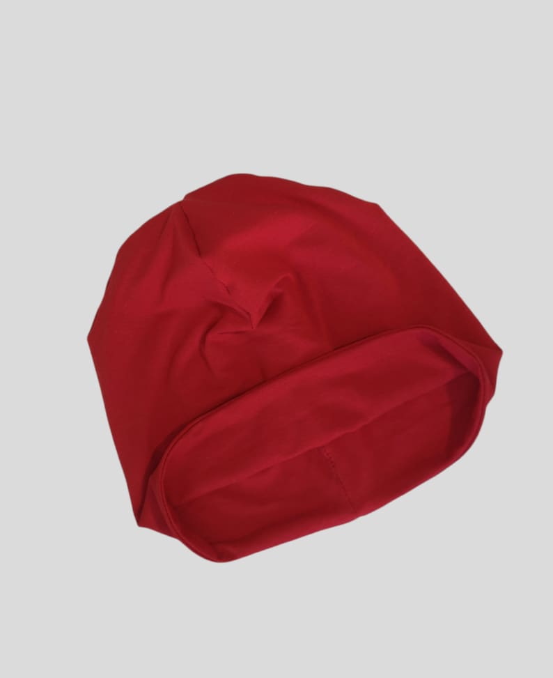 May include: A solid red beanie hat made of soft fabric. The hat is a simple, classic design, with a folded brim and a rounded crown. The hat is shown against a light gray background.