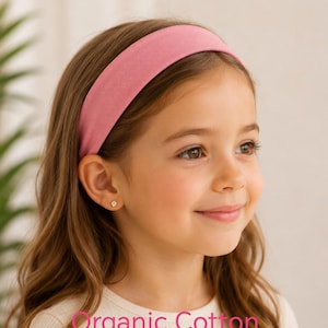 May include: A solid pink headband made of organic cotton. The text on the image reads Organic Cotton 35+ colours. The headband is a wide, fabric-covered band that sits across the top of the head, providing a simple and functional hair accessory.