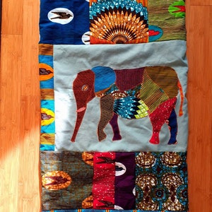 Peut inclure: Une courtepointe en patchwork avec un motif d'éléphant au centre. La courtepointe est faite de différents tissus colorés avec des motifs géométriques et des oiseaux. L'éléphant est fait d'un patchwork de différents tissus, dont du bleu, du rouge, du vert et du jaune.