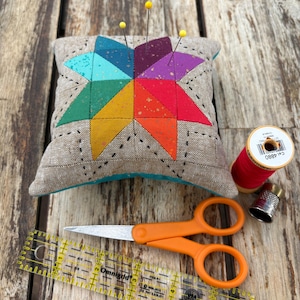 May include: A square pin cushion with a beige linen cover featuring a multicolored star pattern in teal, green, yellow, orange, red, and purple. Three yellow-headed pins are inserted into the top. Includes orange scissors, a red thread spool, a metal thimble, and a clear 5-inch Omnigrid ruler.
