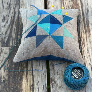 May include: A square pincushion with a geometric design in shades of blue and teal, with yellow pins inserted. A spool of blue thread and needle are next to the pincushion. The pincushion is on a weathered wooden surface.