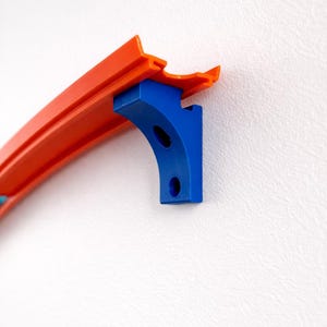 Puede incluir: Primer plano de una pieza de pista de juguete azul y naranja. La pista naranja está sostenida por un soporte azul con dos agujeros. La pista está diseñada para coches de juguete.
