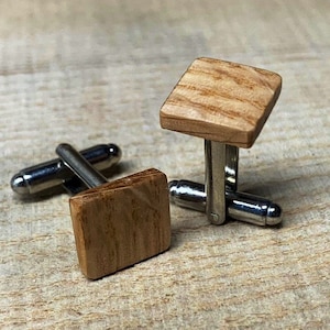 Puede incluir: Un par de gemelos de madera con cierre de metal plateado. La madera es de color marrón claro y tiene un patrón de veta natural.