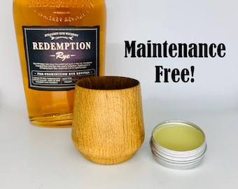 Puede incluir: Una taza de madera, una pequeña lata de bálsamo y una botella de whisky Redemption Rye. El texto "Maintenance Free!" está escrito en letras negras sobre un fondo blanco.