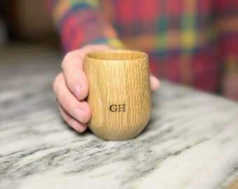 Puede incluir: Una taza de madera con las iniciales "GH" grabadas en el lateral. La taza es de color marrón claro y tiene una veta de madera natural.