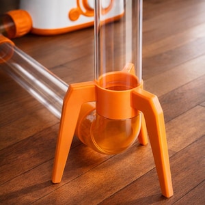 Könnte beinhalten: Ein wissenschaftliches Experimentier-Set aus orangefarbenem und transparentem Kunststoff. Das Set besteht aus einem transparenten Rohr auf einem orangefarbenen Stativfuß, einem weiß-orangenen Gerät und einem transparenten Rohr mit einem orangefarbenen Anschluss. Auf einer Holzoberfläche.