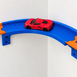 Peut inclure: Une voiture de sport miniature rouge vif est posée sur une section de circuit en plastique bleu incurvée. Le circuit est fixé dans l'angle d'un mur blanc et relié à des segments orange droits, avec des supports de fixation murale.