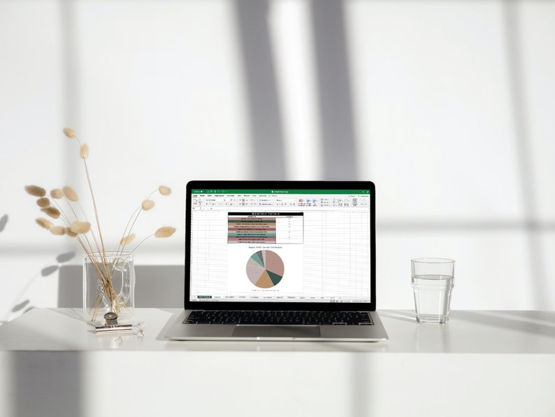 Op de afbeelding: Een laptop met een spreadsheet geopend op het scherm. De spreadsheet bevat een cirkeldiagram met verschillende gekleurde secties. De laptop staat op een wit bureau met een glas water en een vaas met gedroogde bloemen.