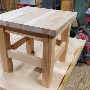 May include: A small, square wooden table with a light-colored top and sturdy, light-brown legs. The table is constructed with visible joinery and has a simple, functional design. The table measures approximately 24 inches square and 18 inches high.