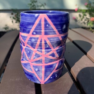 May include: A blue ceramic mug with a pink geometric design. The mug has a textured surface and a wide rim.