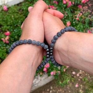 May include: Two matching bracelets made of small, porous black lava stone beads. Each bracelet features a cluster of three smooth, polished, metallic dark grey beads at the centre, creating a uniform and consistent design across both pieces.