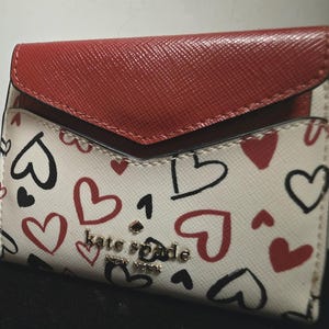 May include: A Kate Spade cardholder with a red flap and a white body. The white body is decorated with red and black heart doodles. The brand name "kate spade new york" is in gold lettering.