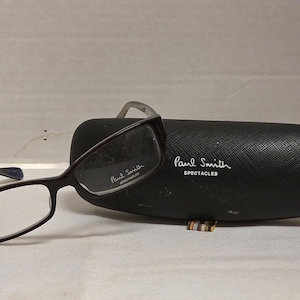 May include: Dark brown rectangular eyeglasses with clear lenses and the text Paul Smith SPECTACLES printed on the right lens. A matching black textured hard shell glasses case is included, featuring the white text Paul Smith SPECTACLES on the front.