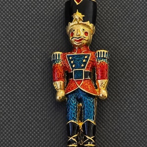 May include: A gold-tone metal brooch shaped like a nutcracker soldier. The figure features a black hat with a red gemstone, a red and blue enameled uniform with gold accents, and black boots. The design includes a textured finish on the enamel.