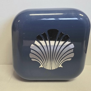 May include: A navy blue, square-shaped container with rounded edges. A silver seashell design is centered on the front. The container has a glossy finish and appears to be made of a solid material.