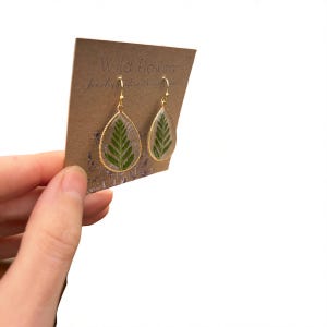 May include: Teardrop-shaped earrings with a gold-colored frame, each containing a preserved green fern leaf. The earrings are displayed on a brown card with the text "Wild Flowear" and "Jewelry made with real plants".