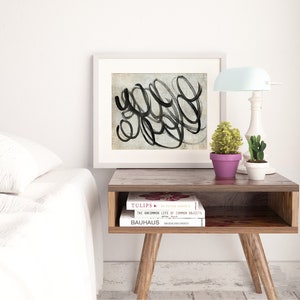 May include: A white framed print with black abstract brushstrokes on a textured background. The print is on a wooden end table with two books, "TULIPS" and "THE UNCOMMON LIFE OF COMMON OBJECTS", and a small potted plant. A white lamp with a light blue shade is on the table.