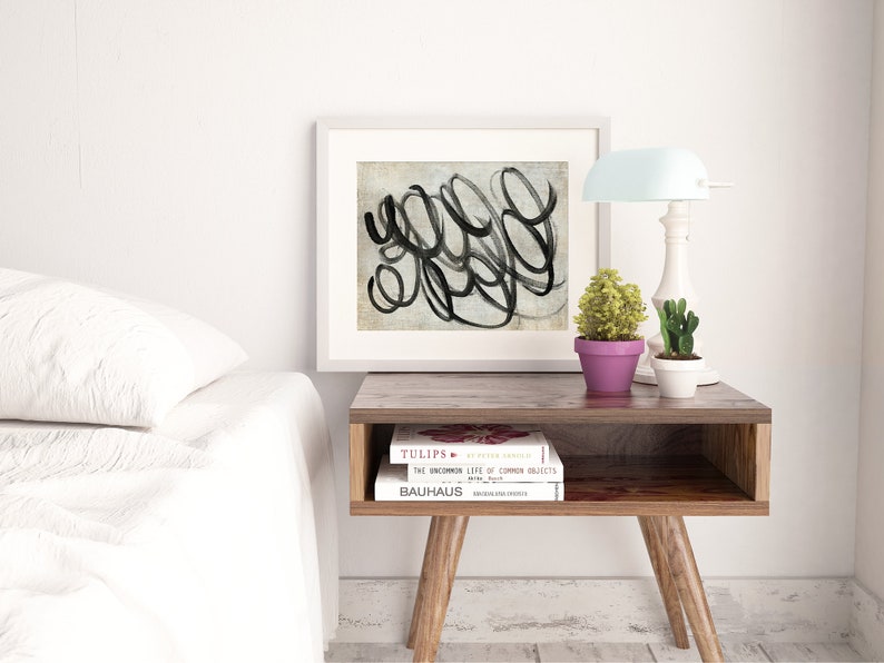 May include: A white framed print with black abstract brushstrokes on a textured background. The print is on a wooden end table with two books, "TULIPS" and "THE UNCOMMON LIFE OF COMMON OBJECTS", and a small potted plant. A white lamp with a light blue shade is on the table.