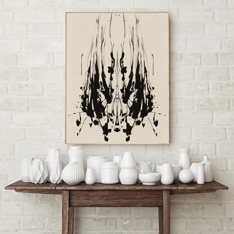 May include: A black and white abstract inkblot print on a beige background. The print is symmetrical and has a dark, organic shape in the center. The print is framed and hanging on a white brick wall above a wooden table with white ceramic vases.