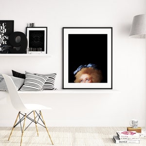 Pu&ograve; includere: Una stampa incorniciata in bianco e nero di un viso di donna con un fiocco blu nei capelli. La donna sta guardando in alto e i suoi occhi sono visibili. La stampa &egrave; appesa a un muro bianco sopra uno scaffale bianco con cuscini a strisce bianche e nere.