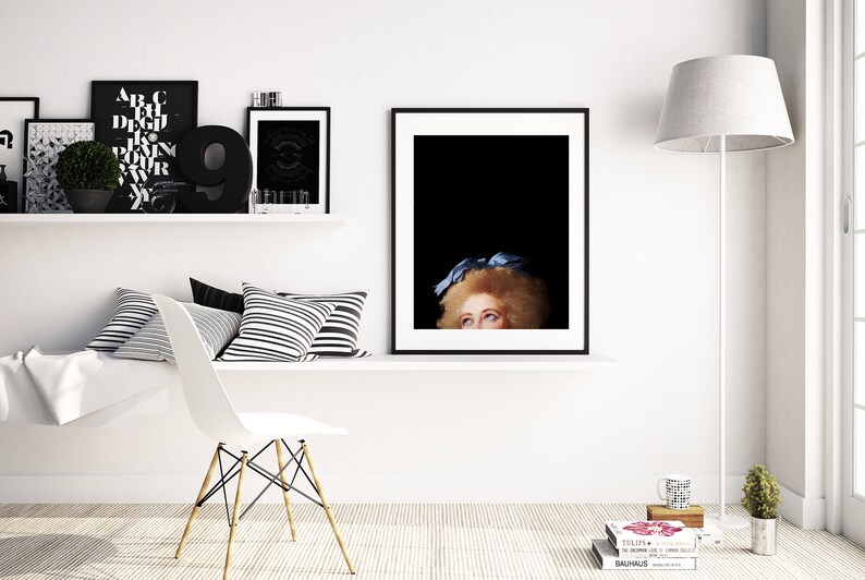 Pu&ograve; includere: Una stampa incorniciata in bianco e nero di un viso di donna con un fiocco blu nei capelli. La donna sta guardando in alto e i suoi occhi sono visibili. La stampa &egrave; appesa a un muro bianco sopra uno scaffale bianco con cuscini a strisce bianche e nere.