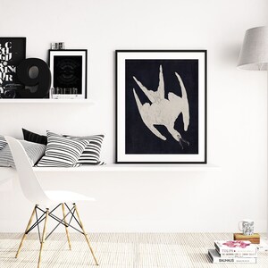 May include: A black and white framed print of a stylized white bird with a long beak against a dark blue background. The print is hanging on a white wall above a white shelf with black and white striped pillows.