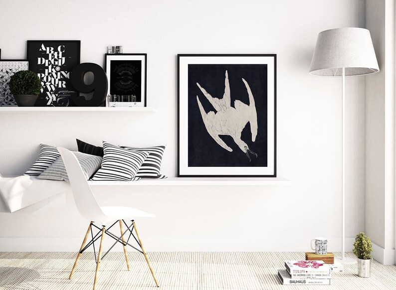 May include: A black and white framed print of a stylized white bird with a long beak against a dark blue background. The print is hanging on a white wall above a white shelf with black and white striped pillows.