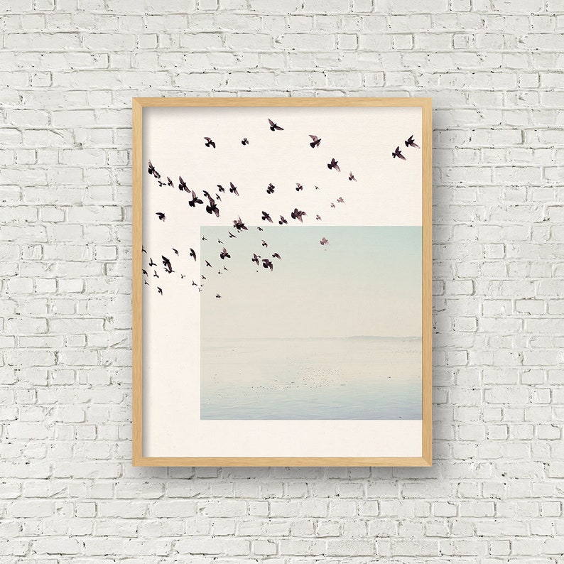 May include: A framed print of a minimalist landscape with a flock of birds flying over a body of water. The sky is a soft blue and the water is a light blue. The birds are black silhouettes against the sky.