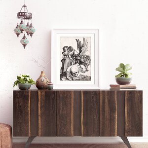 May include: A black and white print of a Renaissance-era engraving depicting a winged figure holding a skull. The print is framed in a white frame and is displayed on a wooden shelf.