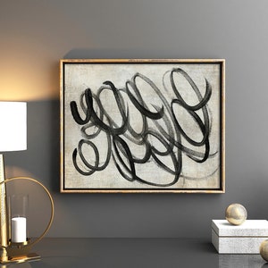 May include: A black and white abstract painting with a gold frame hangs on a gray wall above a gold table lamp, a gold candle holder, and a white vase with a gold design.