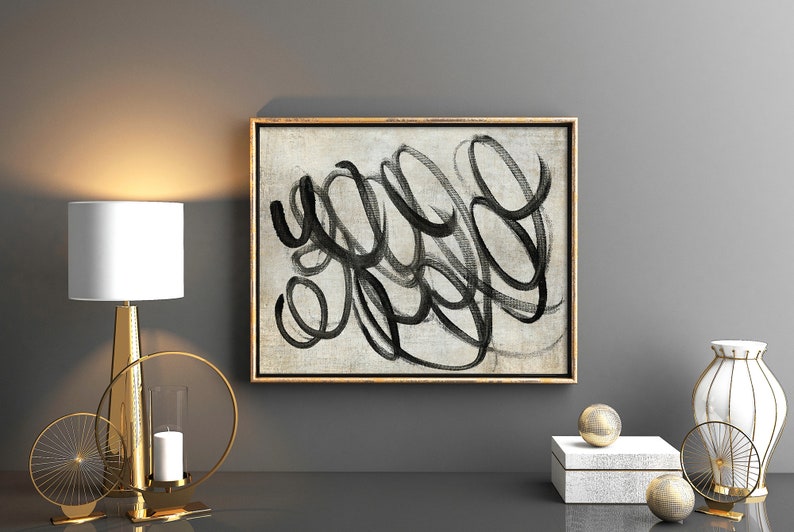 May include: A black and white abstract painting with a gold frame hangs on a gray wall above a gold table lamp, a gold candle holder, and a white vase with a gold design.