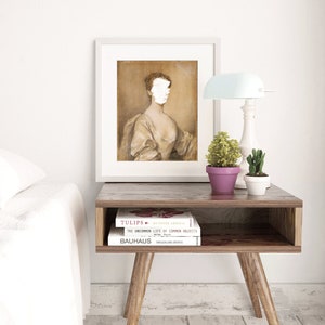 May include: A framed portrait of a woman with a white painted-over face, wearing a brown dress. The portrait is hanging on a white wall above a wooden side table with two books stacked on top of each other. The books are titled "Tulips" and "Bauhaus". A small potted plant sits on the table next to the books.