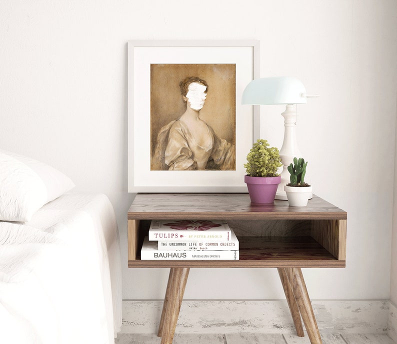 May include: A framed portrait of a woman with a white painted-over face, wearing a brown dress. The portrait is hanging on a white wall above a wooden side table with two books stacked on top of each other. The books are titled "Tulips" and "Bauhaus". A small potted plant sits on the table next to the books.