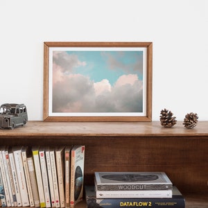May include: A framed photograph of a cloudy sky with a light blue and pink color scheme. The photograph is on a wooden shelf with a small metal car model, two pine cones, and a row of books.