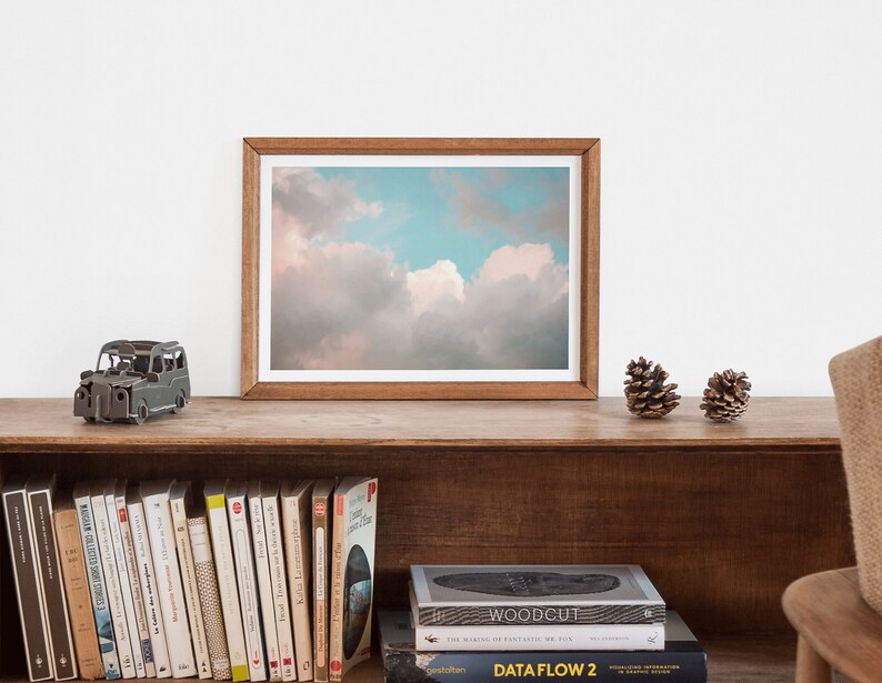 May include: A framed photograph of a cloudy sky with a light blue and pink color scheme. The photograph is on a wooden shelf with a small metal car model, two pine cones, and a row of books.