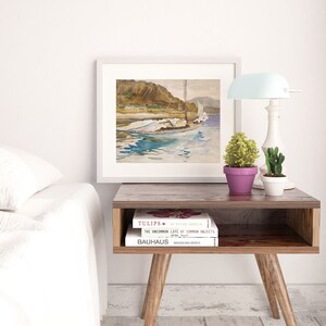May include: A white framed watercolor painting of a sailboat on a body of water with a mountain range in the background. The painting is on a wooden table with two books stacked on top of each other. The books are titled "Tulips" and "The Uncommon Life of Common Objects".