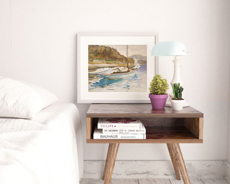 May include: A white framed watercolor painting of a sailboat on a body of water with a mountain range in the background. The painting is on a wooden table with two books stacked on top of each other. The books are titled "Tulips" and "The Uncommon Life of Common Objects".