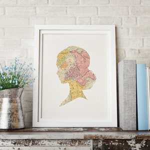 May include: A white framed print of a silhouette of a person's head with a vintage map design. The map is in shades of pink, yellow, and blue.